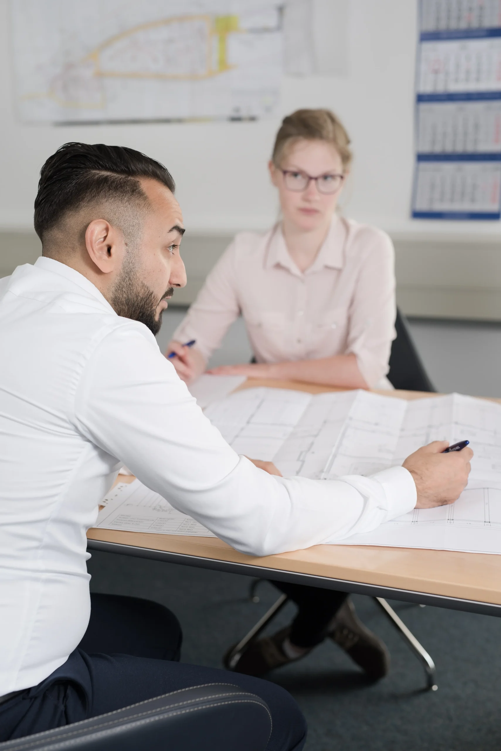 Zwei Personen sitzen an einem Tisch auf dem ein großer Bauplan liegt.